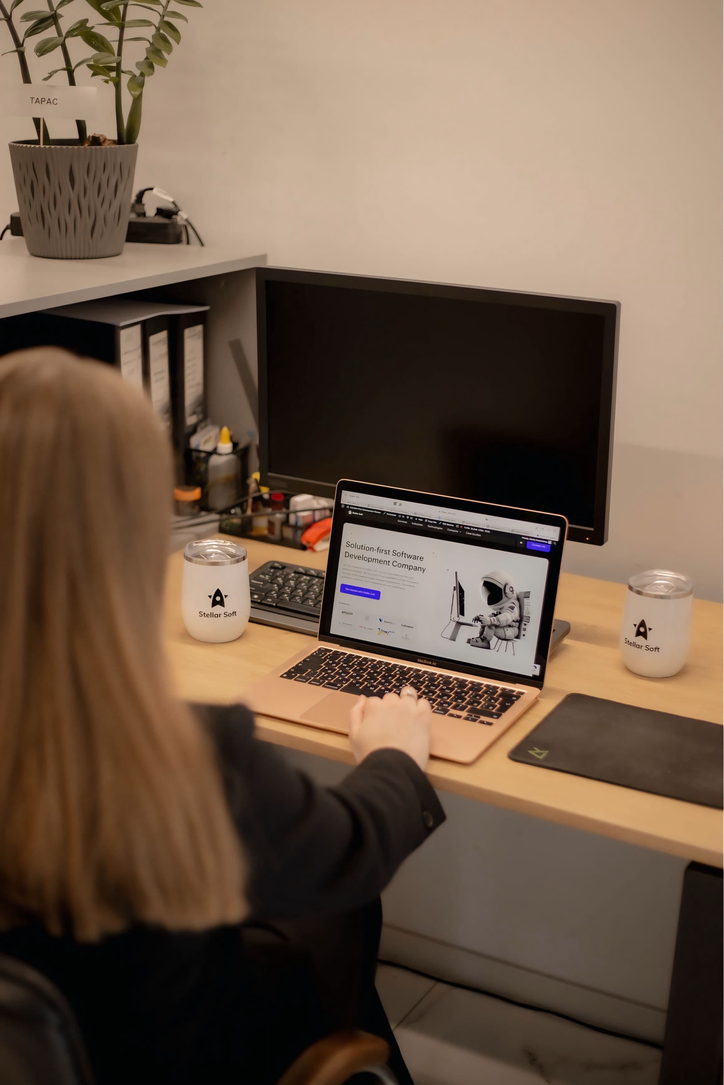 Person using a laptop at a desk with a blank monitor and office supplies.