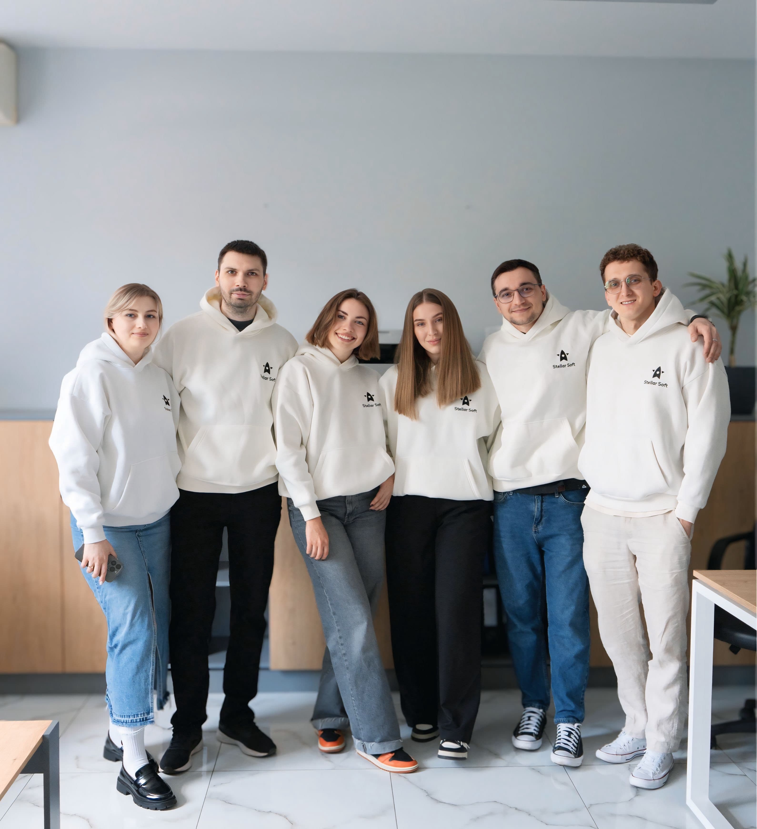 Group of six people wearing white hoodies standing together in a bright room.