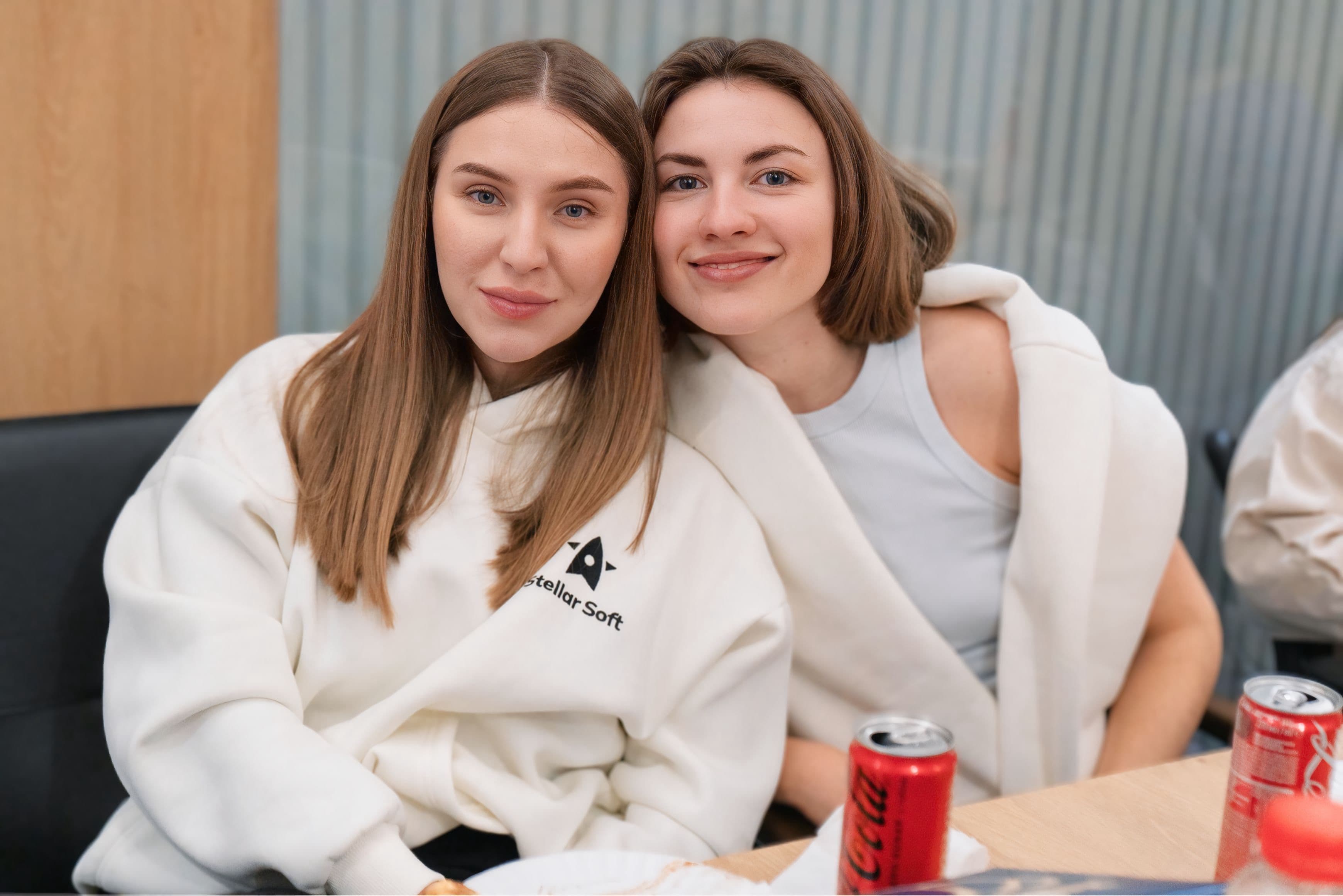 Two women sitting closely, smiling, with soda cans on the table.