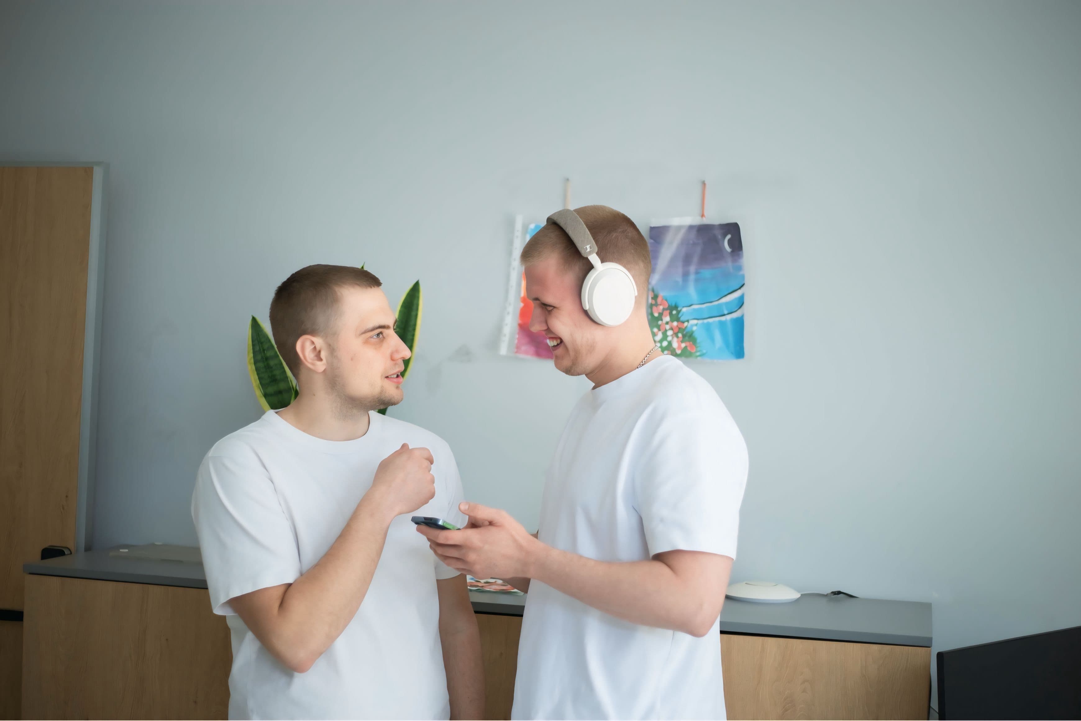 Two men in white shirts talking, one with headphones holding a smartphone.