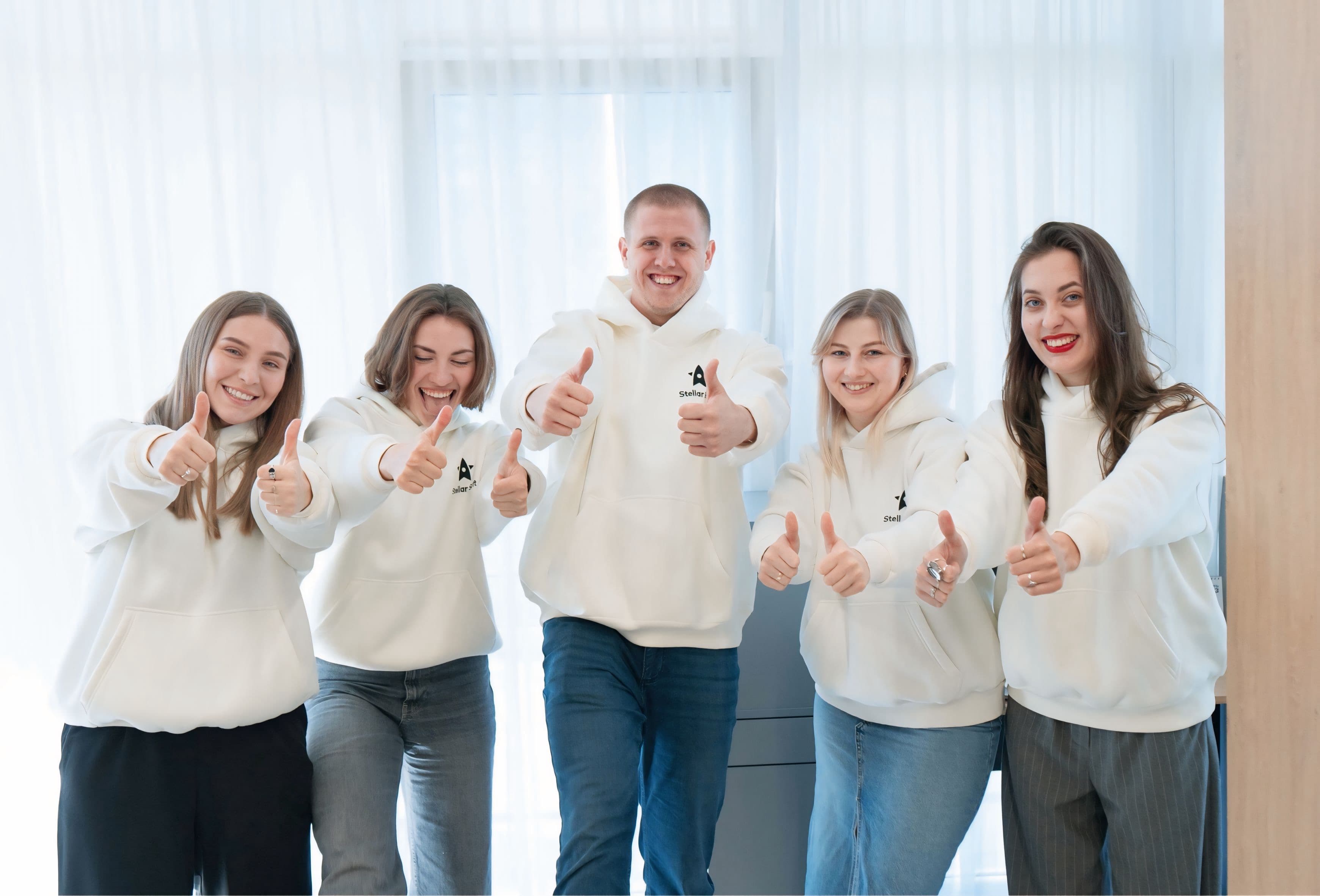 Five smiling people in white hoodies giving thumbs up indoors.