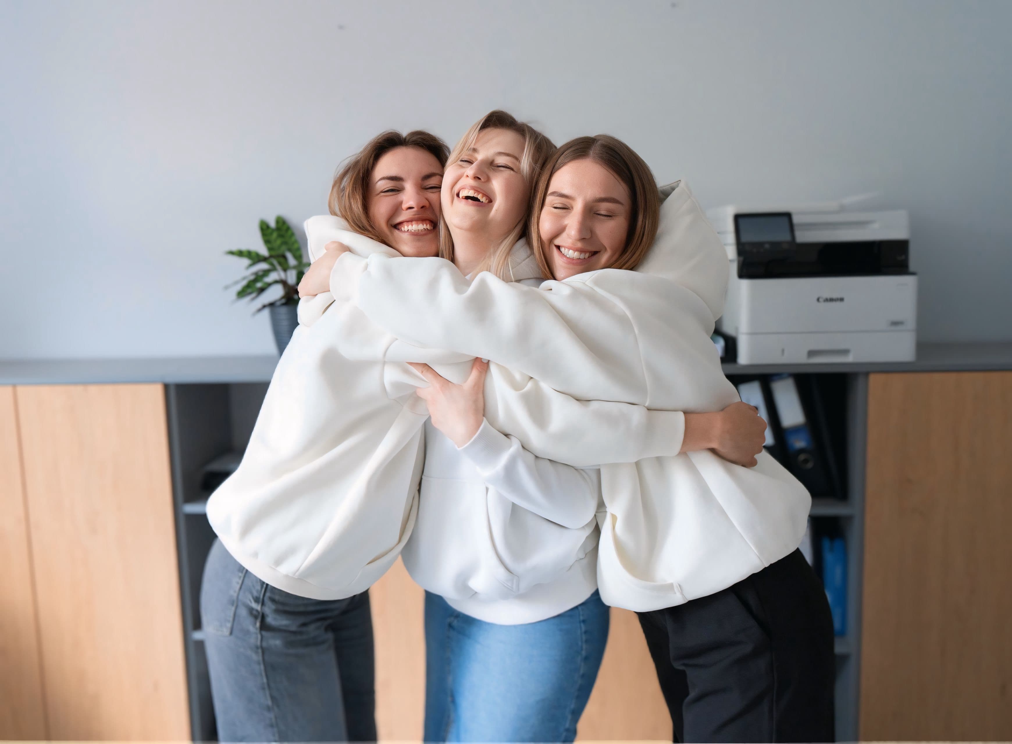 Three people hugging and smiling, wearing white sweatshirts in an office setting.