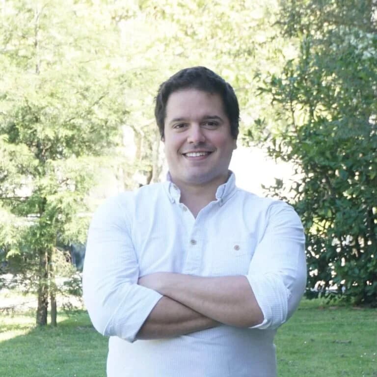 Smiling person in a white shirt standing in a lush green park.