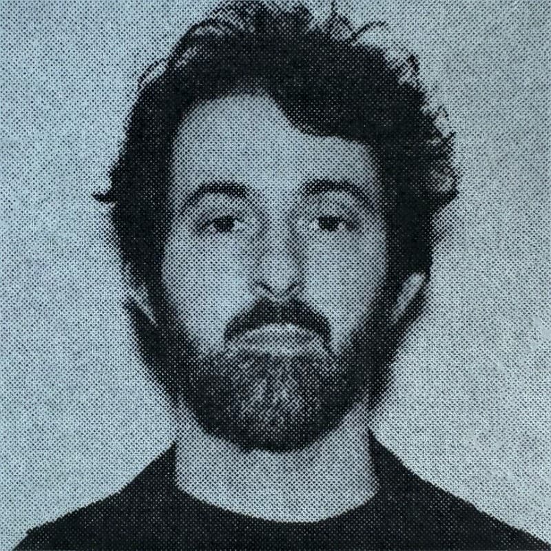 Man with curly hair and beard, wearing a dark shirt, monochrome effect.