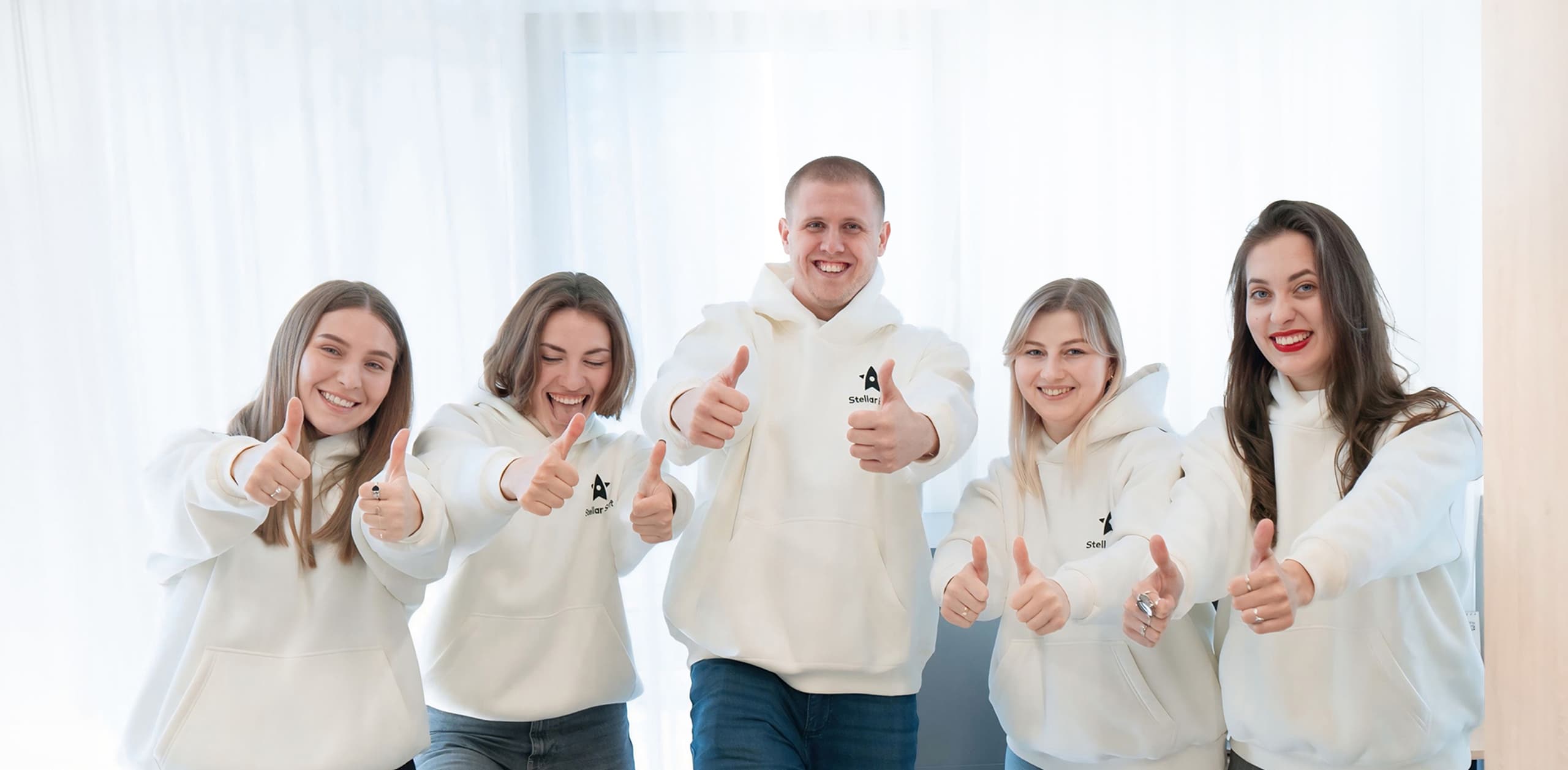 Five people in matching white hoodies giving thumbs up indoors.